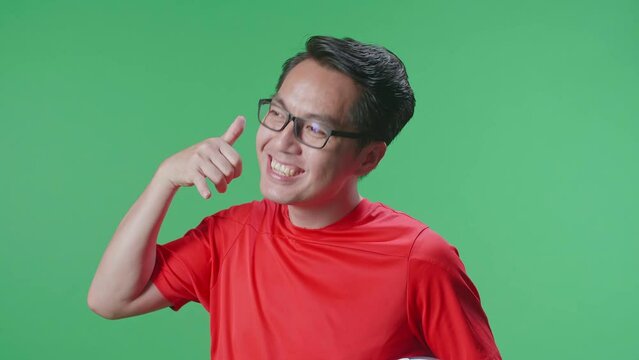 Close Up Side View Of Smiling Asian Man With A Ball Making A Call Me Gesture While Cheering Soccer On Green Screen Background
