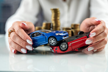 Hands of a woman holding two car models simulating an accident near a mountain of coins.