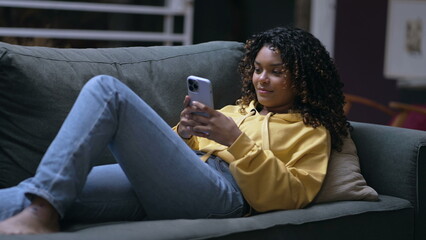 Fototapeta premium One young black woman sitting on couch looking at smartphone device. A relaxed African American 20s girl using cellphone. Person checking social media