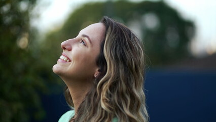 Naklejka premium Happy hopeful young millennial woman standing outside smiling looking at sky with FAITH