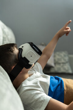 Lateral vertical image of brown boy with virtual reality glasses lying on the sofa raising an arm and pointing a finger to the space created in 3D.