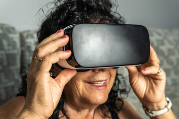 Brunette curly haired older woman wearing a virtual reality goggles and smiling while holding and...