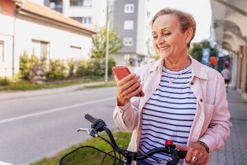 Beautiful happy caucasian active senior woman with bicycle in summer city using smartphone