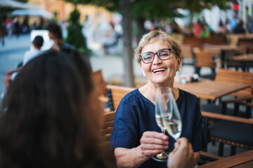 Beautiful happy elderly woman sitting with her friend in a street cafe clinking glasses with...