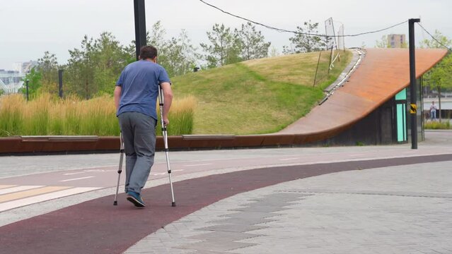 Heavyset millennial man walks on crutches along a track allocated for running in a city park