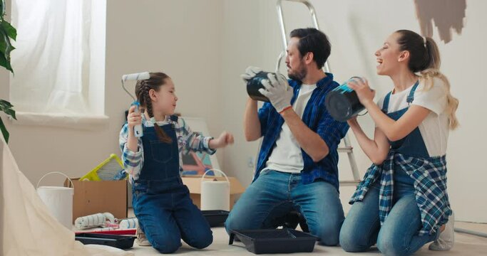 Happy family moved into new house. They sit on floor, hold paint buckets and rollers, dance, smile and sing. Mom, dad and daughter are dressed in jeans and checkered shirts. Behind is ladder and tools
