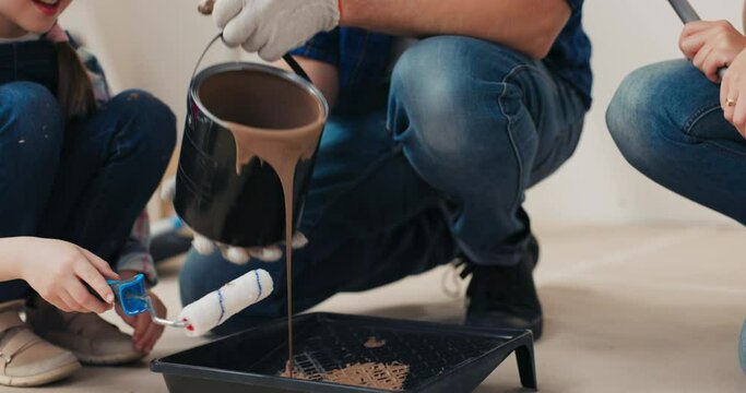 Close-up of man in gloves holding bucket of brown paint and pouring it into cuvette. Daughter in denim overalls is sitting nearby, smiling, holding roller and wiping remains of paint on bucket.