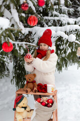 Girl with teedybears and red christmas toys sitting on high chair in the winter forest with toys.