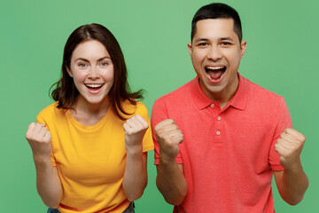 Young fun couple two friends family man woman wear basic t-shirts together doing winner gesture celebrate clenching fists say yes isolated on pastel plain light green color background studio portrait.