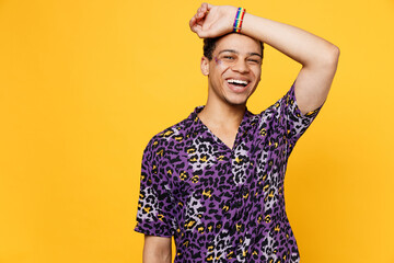 Young gay man wearing purple animal print shirt put hand on forehead show rainbow flag bracelet on arm isolated on bright plain yellow color background studio portrait. Lifestyle lgbtq pride concept.