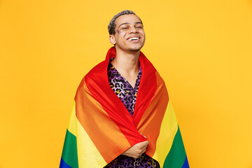 Young smiling cheerful happy gay man wearing purple animal print shirt wrapped in colorful striped rainbow flag isolated on bright plain yellow color background studio. Lifestyle lgbtq pride concept.
