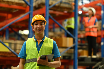 Portrait of male warehouse worker wearing safety hardhat and vest holding digital tablet standing in large retail warehouse