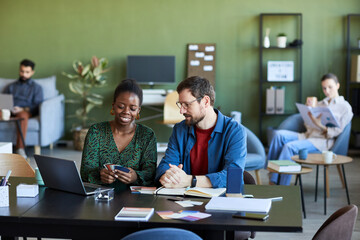 Happy young female designer showing color palette to male colleague whlle both sitting by workplace and preparing new project