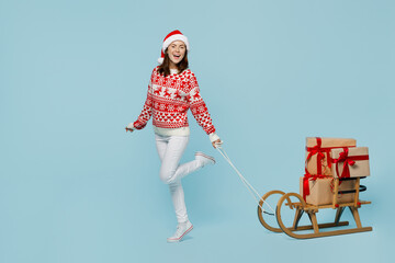 Full body fun side view young merry woman wear red knitted sweater Santa hat posing carrying gifts on a sled go isolated on plain pastel light blue cyan background Happy New Year 2023 holiday concept