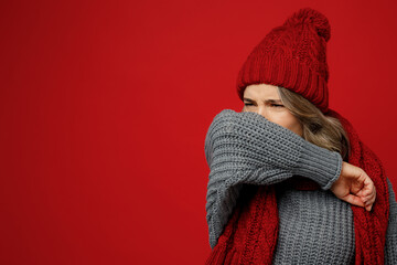 Young sad sick woman with runny nose wear warm gray sweater scarf hat sneeze into sleeve isolated on plain red background studio portrait Healthy lifestyle ill disease treatment cold season concept