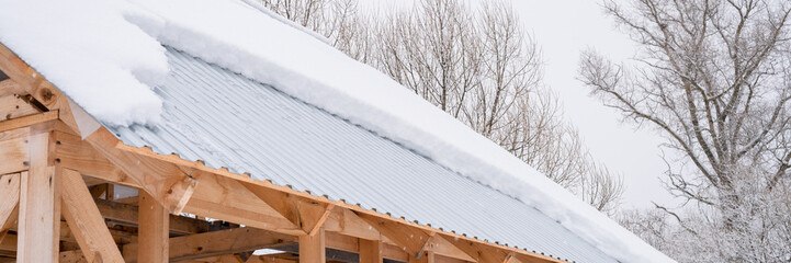 snow sliding down from roof. building construction house with metal roof covered fresh icy frozen snow and snowflakes on frosty winter day in country village suburb. snowy winter season. banner