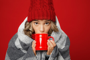 Close up young woman wear scarf hat wrapped in grey plaid drink sip hot tea milk look camera isolated on plain red background studio Healthy lifestyle ill sick disease treatment cold season concept