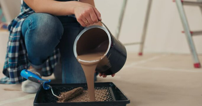 Close-up of girl's hands holding bucket of brown paint and pouring it in cuvette. Woman puts bucket on side, takes roller and dips it in paint. Girl in denim overalls is ready to paint walls.