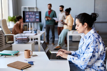 Side view of young serious female employee typing on laptop keypad while sitting by desk in openspace office against her colleagues