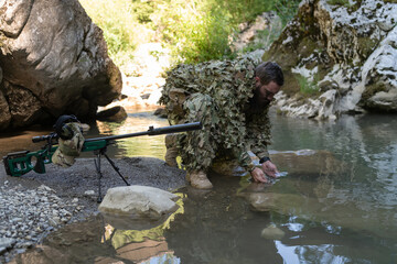 Soldier in a camouflage suit uniform drinking fresh water from the river. Military sniper rifle on the side. 