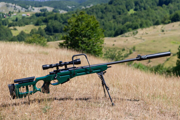 Fototapeta na wymiar A green military sniper rifle with a scope for long distance tactical modern warfare in yellow grass blue sky