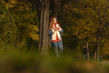 Young woman with dreadlocks in warm clothes standing near tree and drinking coffee. Female in casual outfit standing near green trees and drinks hot coffee or tea while resting in park on sunny day