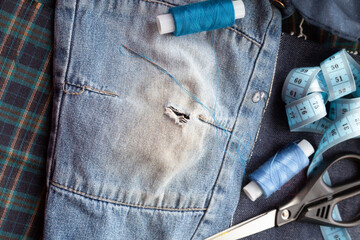 Clothing repair. blue jeans with a hole, scissors, darning thread close-up - obrazy, fototapety, plakaty