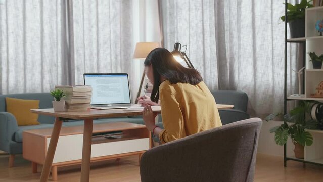 Back View Of A Young Female Copy The Text On A Laptop To A Book While Studying At Home

