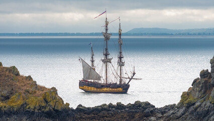Sailing ship on the blue waters of the atlantic ocean on the coastline of Brittany, France, close to the village of Cancale. Rocks at both sides. Dramatic sky with clouds on the background.