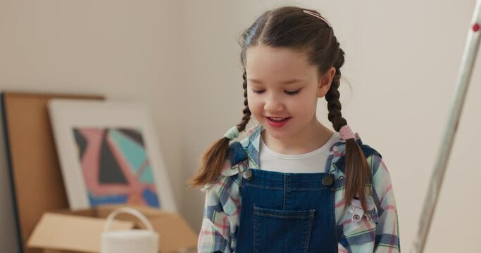 Close-up of smiling girl with pigtails holding roller in hands and dipping it in brown paint. Seven-year-old girl is wearing denim overalls and checkered shirt. Behind are boxes, cans of paint.