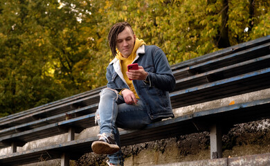 Fototapeta premium Young man with dreadlocks browsing mobile phone, sitting on old shabby bench of street bleacher.