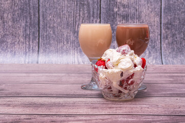 On wooden table are a vase filled ripe strawberries and covered cream, and two glasses cocktails. Still life with positive emotions. Festive mood.