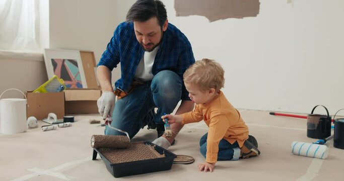 Dad and son are sitting on floor in new apartment. They poured brown paint and dipped roller and brush in it. Man with beard in blue shirt and gloves and son in orange sweater get up and paint walls.