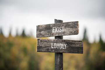 vintage and rustic wooden signpost with the weathered text quote exist loudly, outdoors in nature. blurred out forest fall colors in the background.