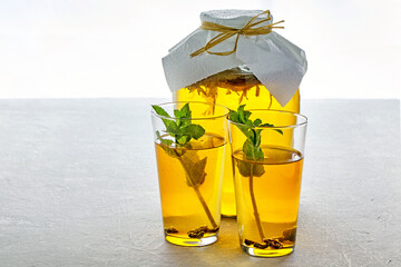 A jar of kombucha and two glass cups of kombucha are decorated with mint on a light gray concrete...