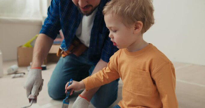 Dad and son are painting walls in new house. They poured brown paint and dipped the roller and brush in it. Man with beard in blue shirt and gloves shows son in orange sweater how to do it right.