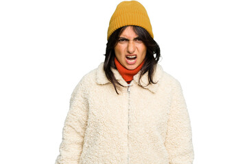 Young Indian woman wearing winter jacket and a wool cap isolated screaming very angry and aggressive.