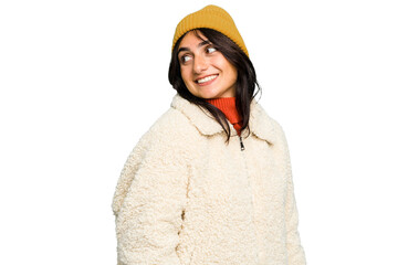 Young Indian woman wearing winter jacket and a wool cap isolated looks aside smiling, cheerful and...
