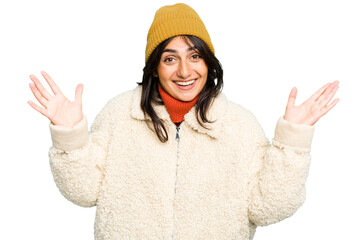 Young Indian woman wearing winter jacket and a wool cap isolated receiving a pleasant surprise, excited and raising hands.