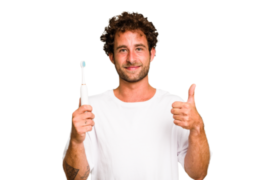 Young caucasian man holding an electric toothbrush isolated smiling and raising thumb up