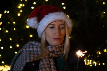 Sad young woman in a Santa Claus hat looks at a sparkler in her hands against the background of Christmas decorations of the city at night. Focus on the sparkler