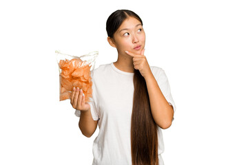 Young asian woman holding crips isolated on green chroma background looking sideways with doubtful and skeptical expression.