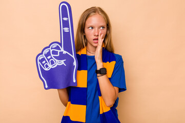 Little caucasian sports fan girl isolated on beige background is saying a secret hot braking news and looking aside