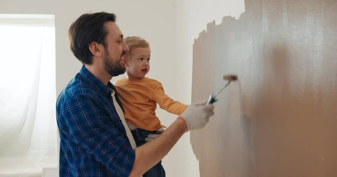 Dad in blue checkered shirt holds son in orange sweater. They hold roller in hands and paint the wall in brown. Bearded man in work gloves talks to son and smiles. Sun is shining outside the window.