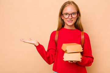 Little caucasian student girl holding burgers isolated on beige background showing a copy space on a palm and holding another hand on waist.