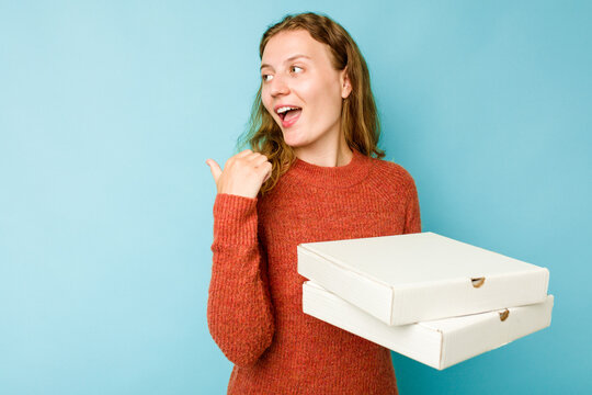 Young caucasian woman holding pizzas boxes isolated on blue background points with thumb finger away, laughing and carefree.