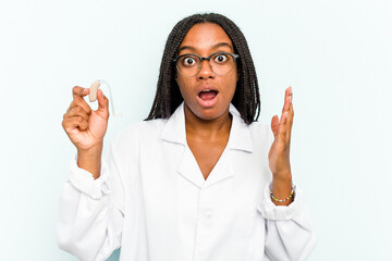 Young African American otorhinolaryngologist woman holding a hearing aid isolated on blue background surprised and shocked.