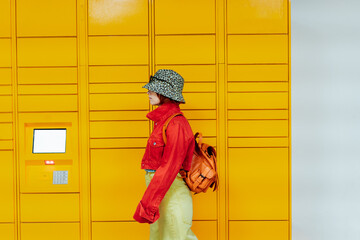 Hipster fashion young woman in bright clothes going past modern postal automatic mail terminal with...