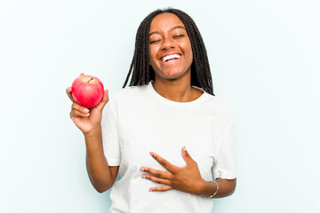 Young African American woman holding an apple isolated on blue background laughs out loudly keeping hand on chest.