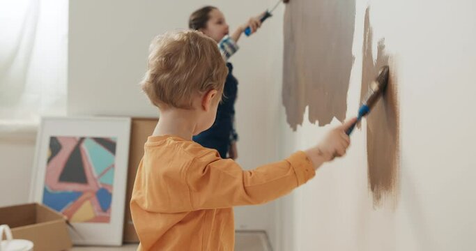 Two-year-old boy in orange sweater holds brush in hands and paints the wall in brown. Boy has blond hair and looks at sister. Girl with pigtails looks at brother and smiles. There is painting behind.